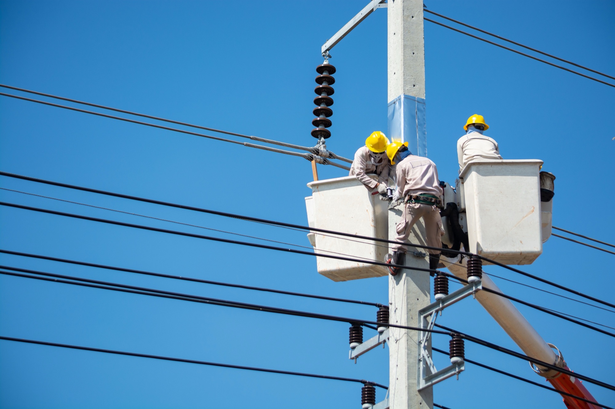 Electricians working on overhead power lines from bucket lifts against a clear sky